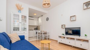 A modern living room with a blue sofa, a TV, and a small yellow table. There is a kitchen in the background with white cabinets and a dining area.