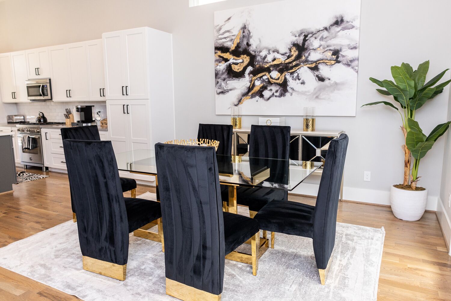 A modern dining area with a glass table and black velvet chairs, white cabinetry, and a large abstract painting.
