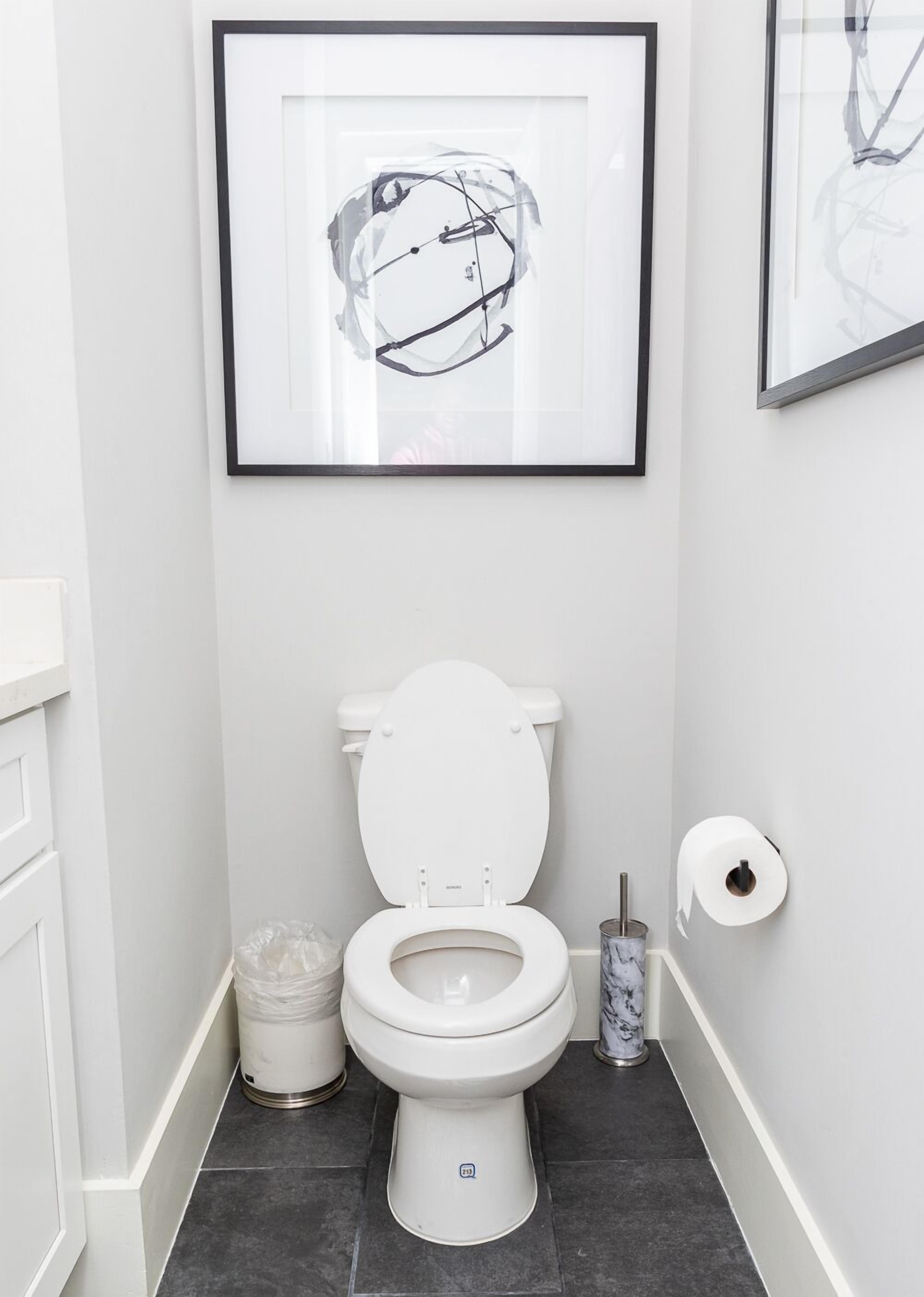 A modern bathroom with a white toilet, a framed abstract artwork above it, and a marble toilet brush holder.