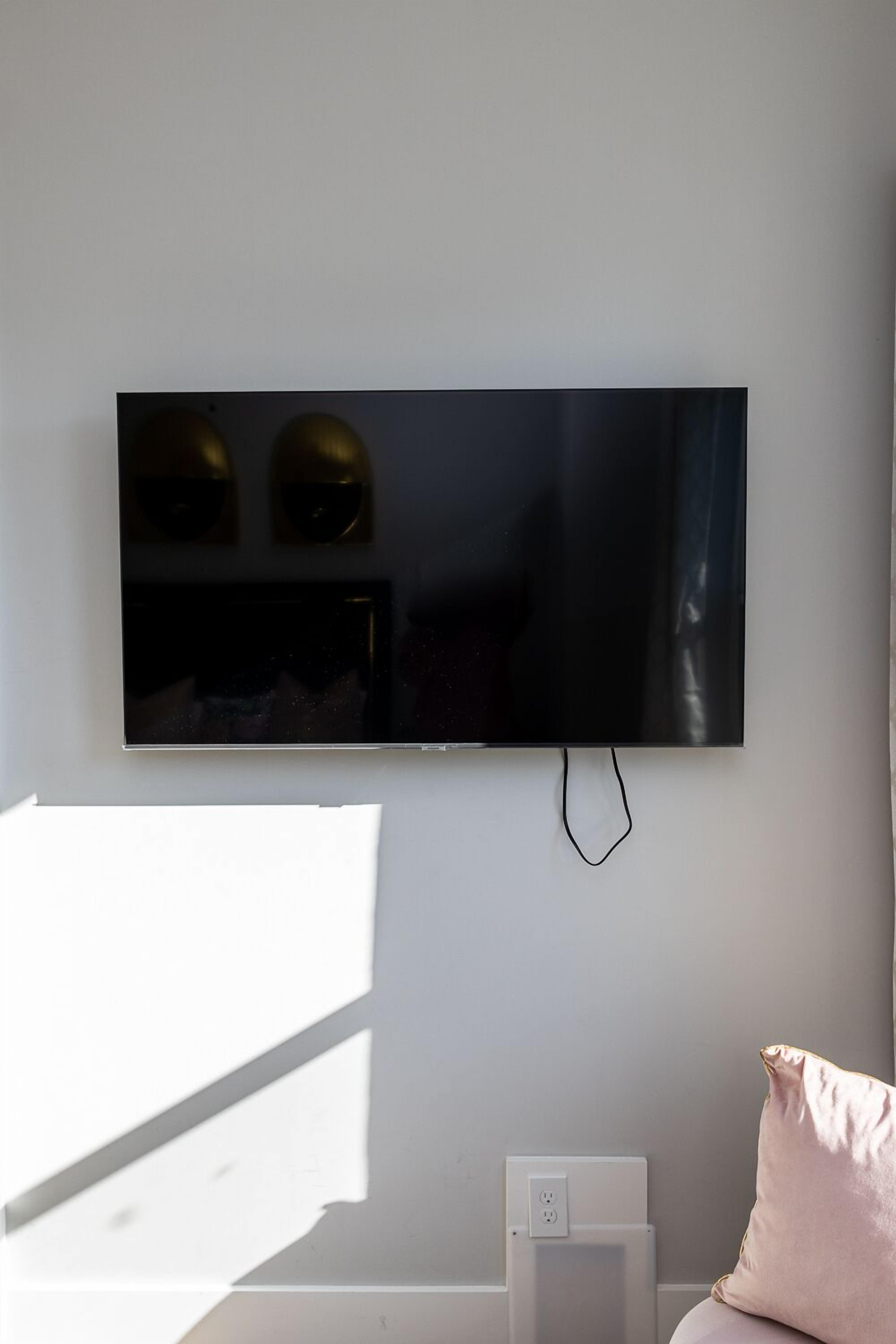 Una habitación moderna con un televisor de pantalla plana grande montado en la pared, una mesita de noche blanca y una almohada rosa.
