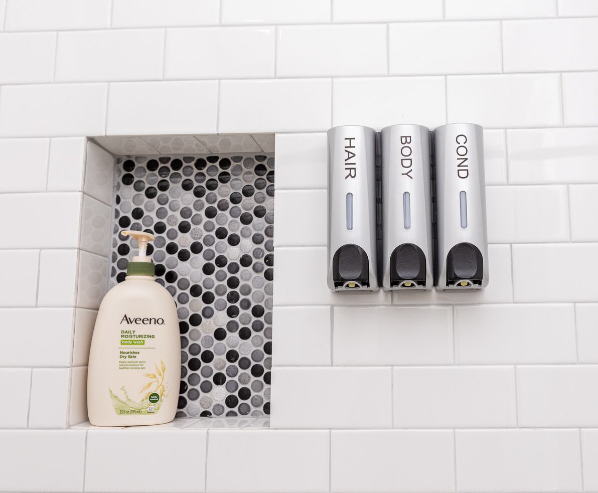 Een badkamer met een witte betegelde muur, een fles Aveeno hydraterende lotion, en drie dispensers met de aanduiding 'HAAR', 'LICHAAM' en 'CONDITIONER'.
