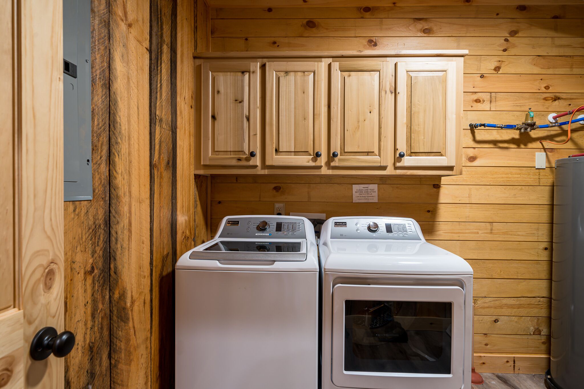 A laundry room with two front-loading washing machines, wooden cabinets above, and a visible electrical panel.