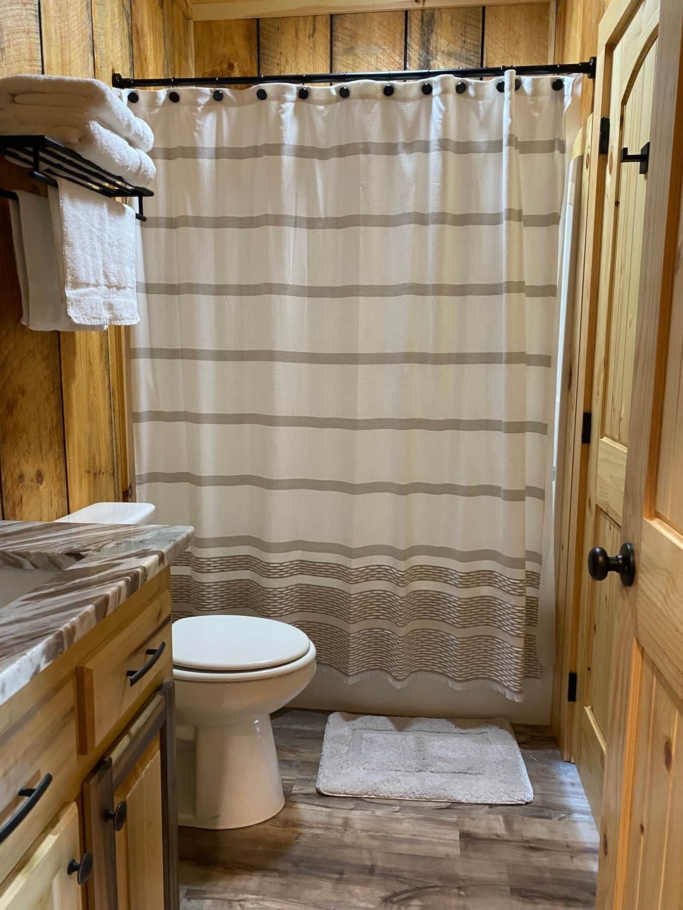 A bathroom with a striped shower curtain, a toilet, a wooden vanity with a marble countertop, and a wooden door.