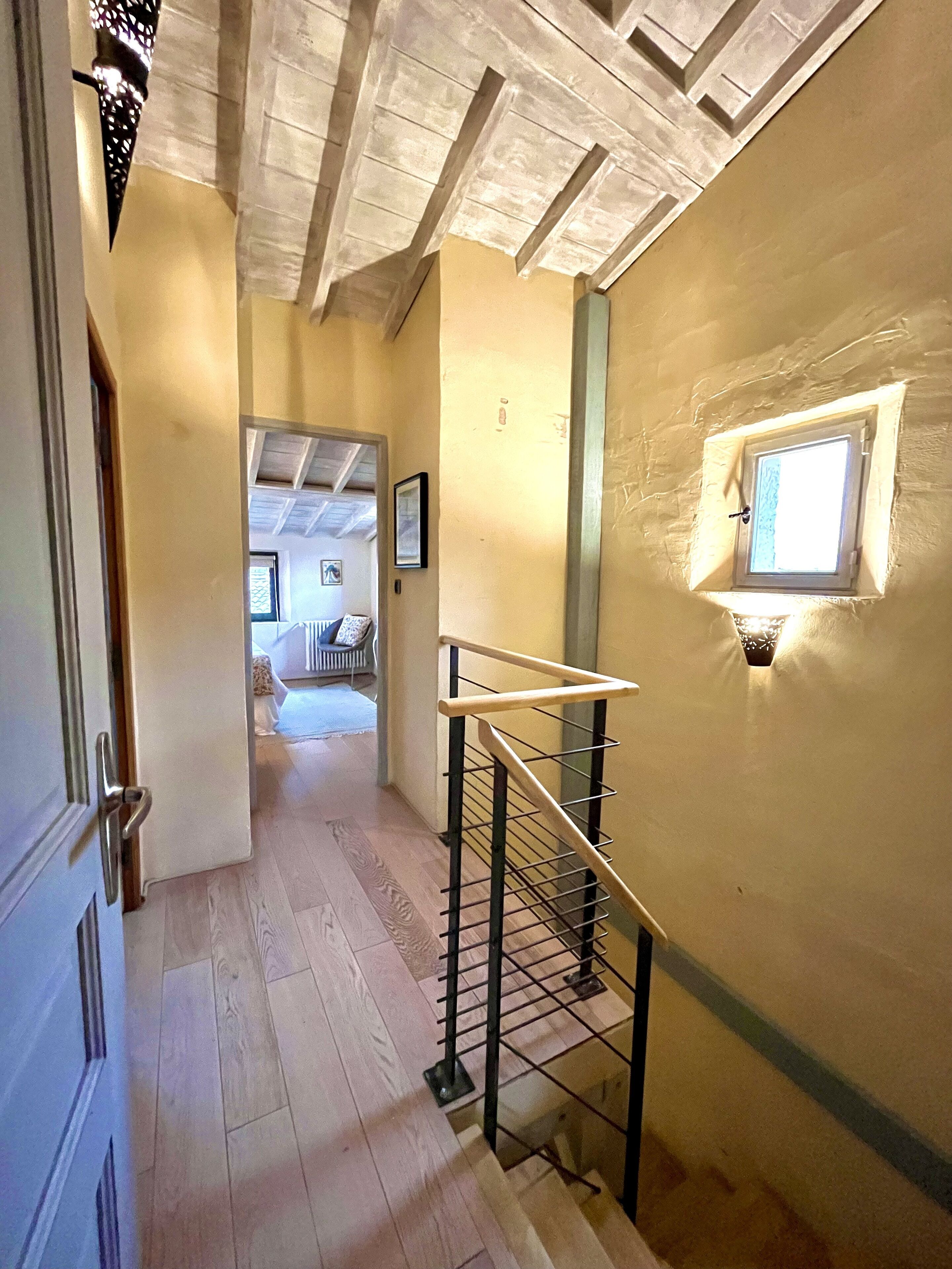 Un couloir doté d’un plancher en bois, d’un escalier avec une rampe en métal et d’une petite fenêtre avec un luminaire.