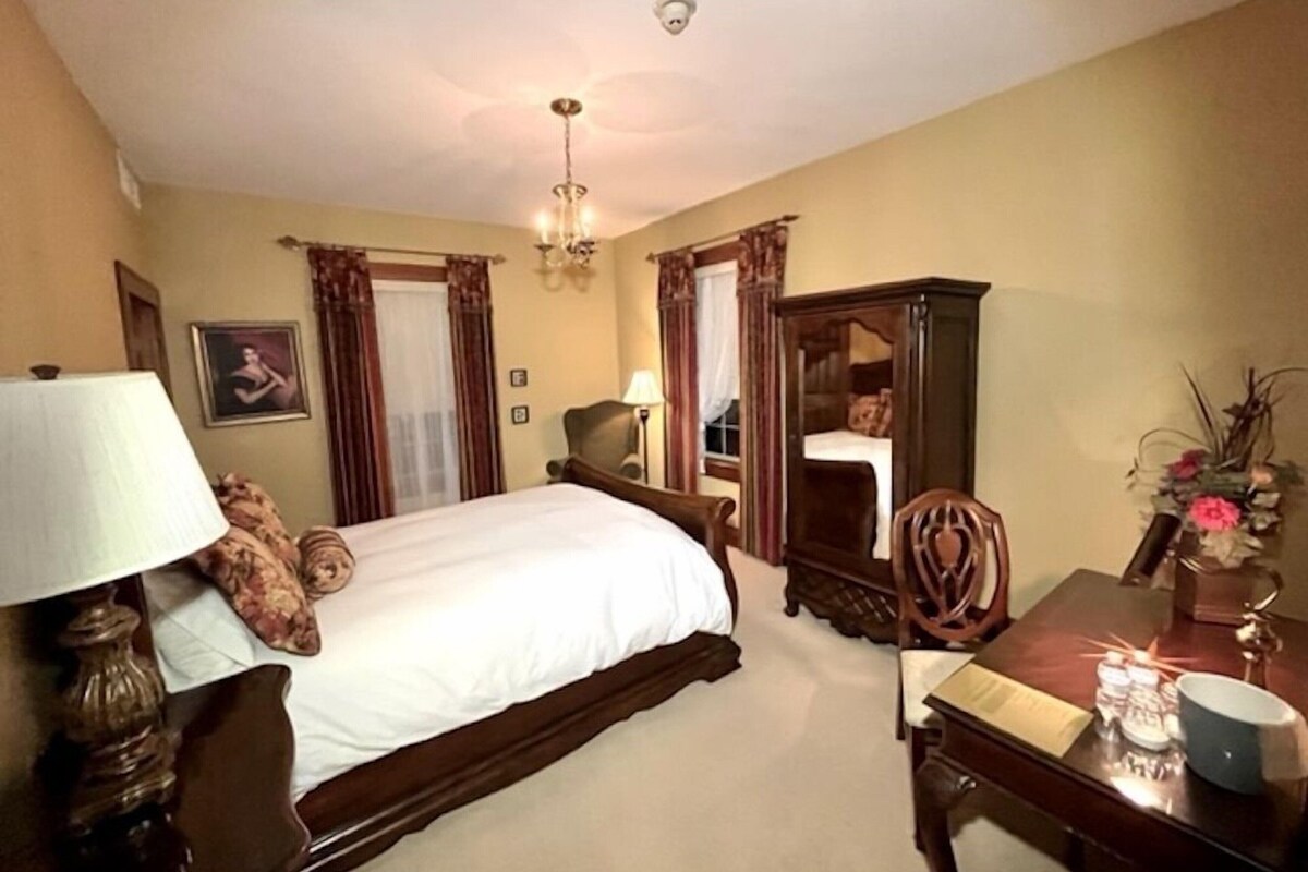 A hotel room with a large bed, a desk, a chair, a wardrobe, and a chandelier.