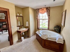 A bathroom with a large bathtub, a vanity with a mirror, and a window with curtains. - The Port Hotel (Port Washington)