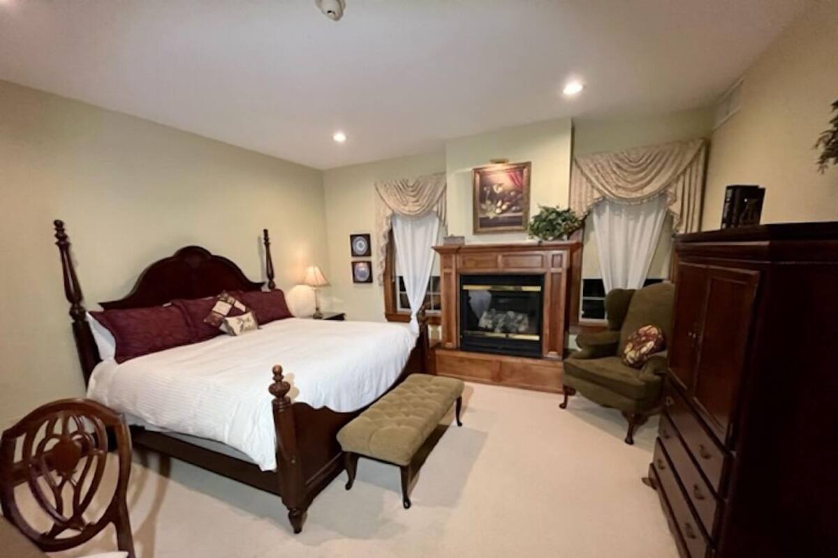 A bedroom with a large bed, a wooden chair, a small table, a fireplace, and a cabinet.