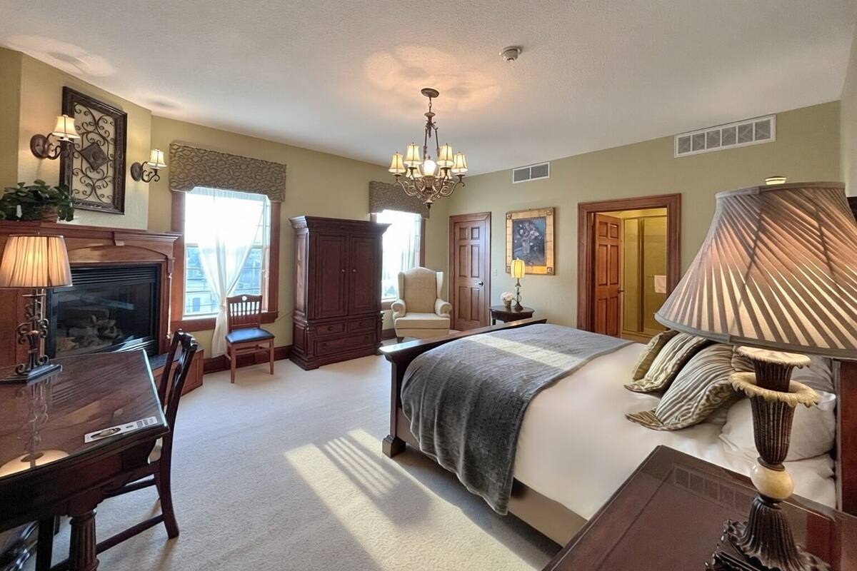 A bedroom with a bed, a desk, a chair, a fireplace, and a chandelier.