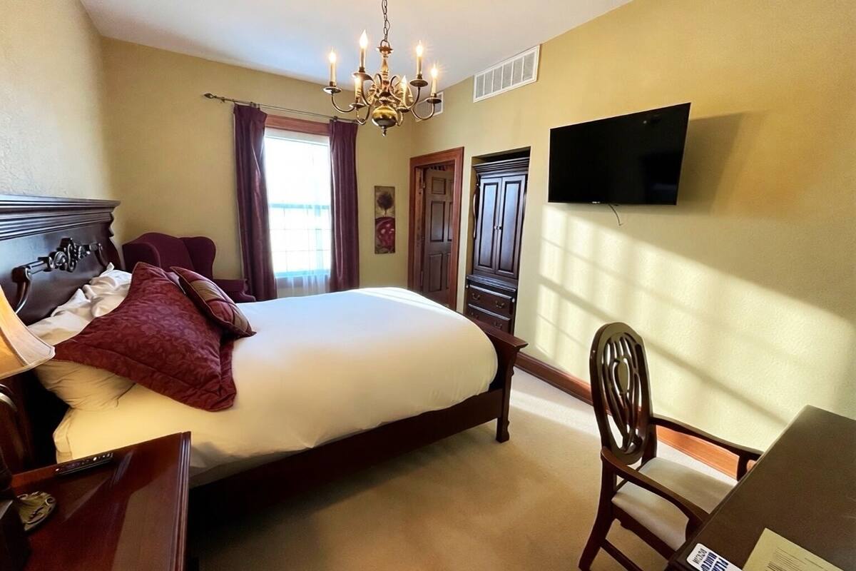 A hotel room with a large bed, a desk with a chair, a television mounted on the wall, and a chandelier.