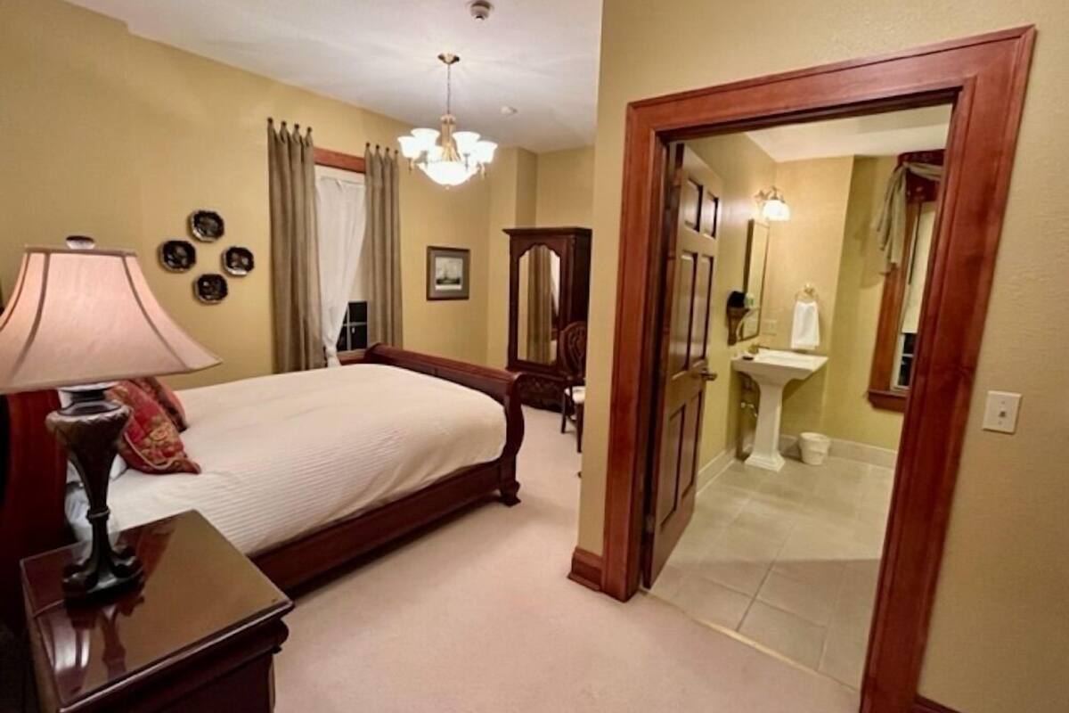 A bedroom with a bed, a nightstand, a lamp, a chandelier, and a bathroom visible through an open door.