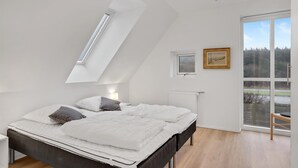A bedroom with a sloped ceiling, a bed with white bedding, a bedside table with a lamp, a window with a view of trees, and a small picture frame on the wall.