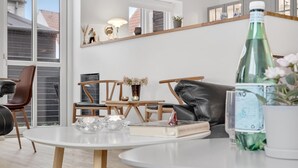 A modern living room with a round white table, two wooden chairs, a black leather sofa, and a large window with a view of buildings.