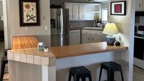 Uma cozinha com ilha de madeira, banquetas pretas, uma geladeira e um abajur.