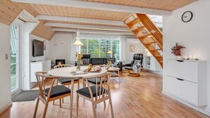 A modern loft with a dining area, kitchen, and living space, featuring wooden beams, large windows, and a staircase.