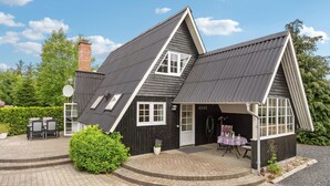 A two-story house with a steeply pitched roof, a chimney, and a small porch with a table and chairs.
