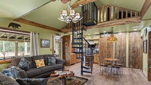 A spacious living area with a large sofa, a dining table with chairs, a staircase, and wooden walls. - Jim Thorpe Rustic Cabin With Hot Tub, EV Charger, BBQ Grill & Outdoor Games! (Jim Thorpe)