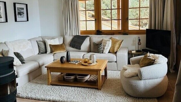 Un salón con sofá, sillón, mesa de centro, televisor y ventanas grandes con cortinas.