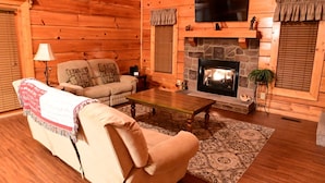 A cozy log cabin interior with a stone fireplace, a sofa, a coffee table, and a television mounted above the fireplace. - SMOKY MOUNTAIN HIGH GETAWAY (Sevierville)