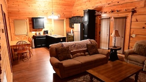 A cozy cabin interior with a wooden dining table, a sofa with a 'Friends' blanket, a kitchen area with a refrigerator, and a lamp on a side table. - SMOKY MOUNTAIN HIGH GETAWAY (Sevierville)