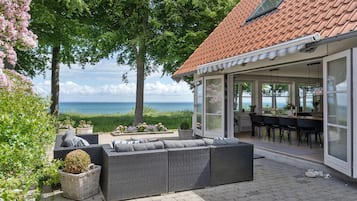 Et overdækket udendørs siddeområde med udsigt til havet, et spiseområde med bord og stole, samt en stor fletsofa.