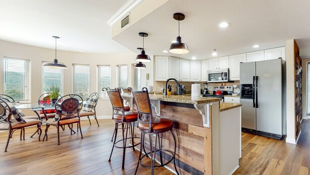 A modern kitchen with wooden flooring, a central island, bar stools, and a dining area with chairs.