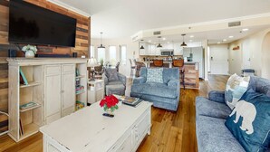 A modern living room with a flat-screen TV, a coffee table with books and a vase of flowers, two sofas, and a kitchen in the background.
