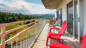 A balcony with a view of a green landscape and mountains, furnished with red chairs and a table.