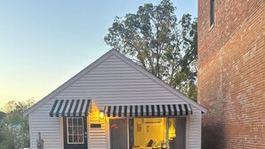 A small, single-story building with a black door, a window with a view of the interior, and a small porch area with two chairs.