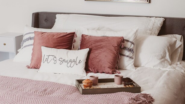 A neatly made bed with a pink and white color scheme, a tray with muffins, and a wall art piece above.
