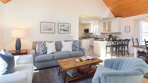 Ein modernes Wohnzimmer mit einem grauen Sofa, einem Holztisch und einem blauen Sessel. Im Hintergrund ist eine Küche mit weißen Schränken und einer Bar erkennbar.