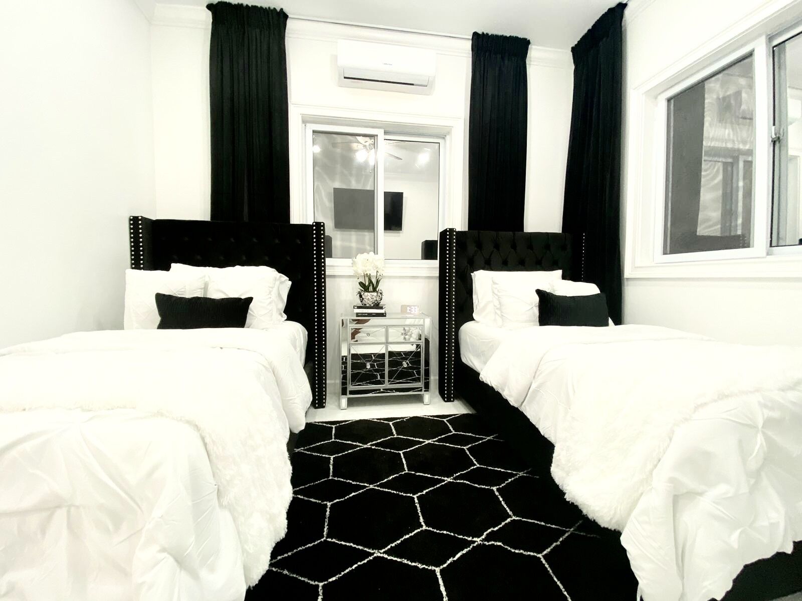 A bedroom with two beds, black and white bedding, a black and white hexagonal rug, and black curtains.