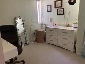 Um quarto com uma escrivaninha, uma cadeira, uma cômoda, um abajur e decorações nas paredes, incluindo quadros e um disco de vinil.