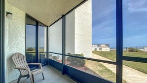 A covered balcony with a chair, large windows, and a view of a building and grassy area.