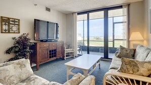 A living room with a television, a coffee table, and a sofa. There is a large window with a view of the outdoors.