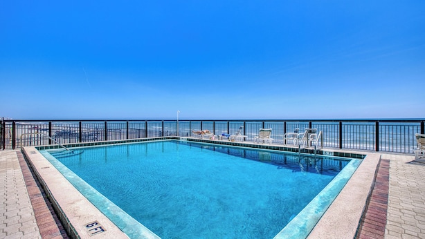 Une piscine rectangulaire à l’eau d’un bleu limpide, entourée d’une clôture en métal noir, avec une vue sur l’océan en arrière-plan.