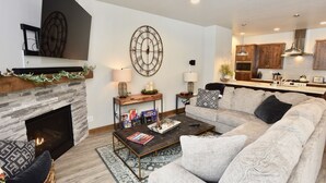 A living room with a fireplace, a large wall clock, a sofa, a coffee table, and a kitchen area in the background. - Silverpine Gem - Downtown - Walk to shops, restaurants, Payette Lake - WiFi (McCall)