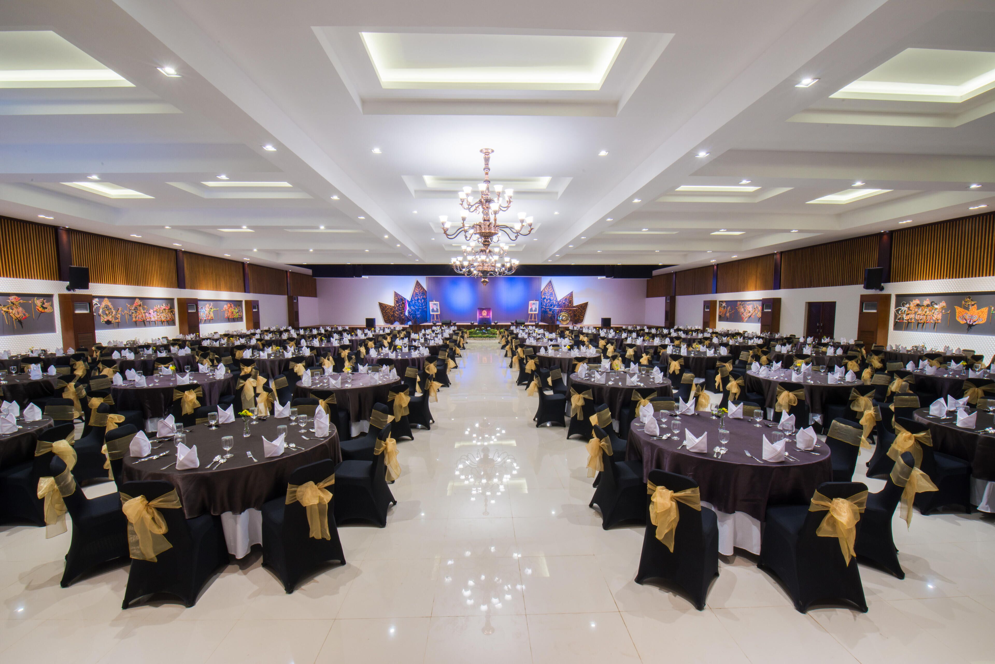 A large banquet hall with round tables and chairs, a chandelier, and a stage with a blue backdrop.