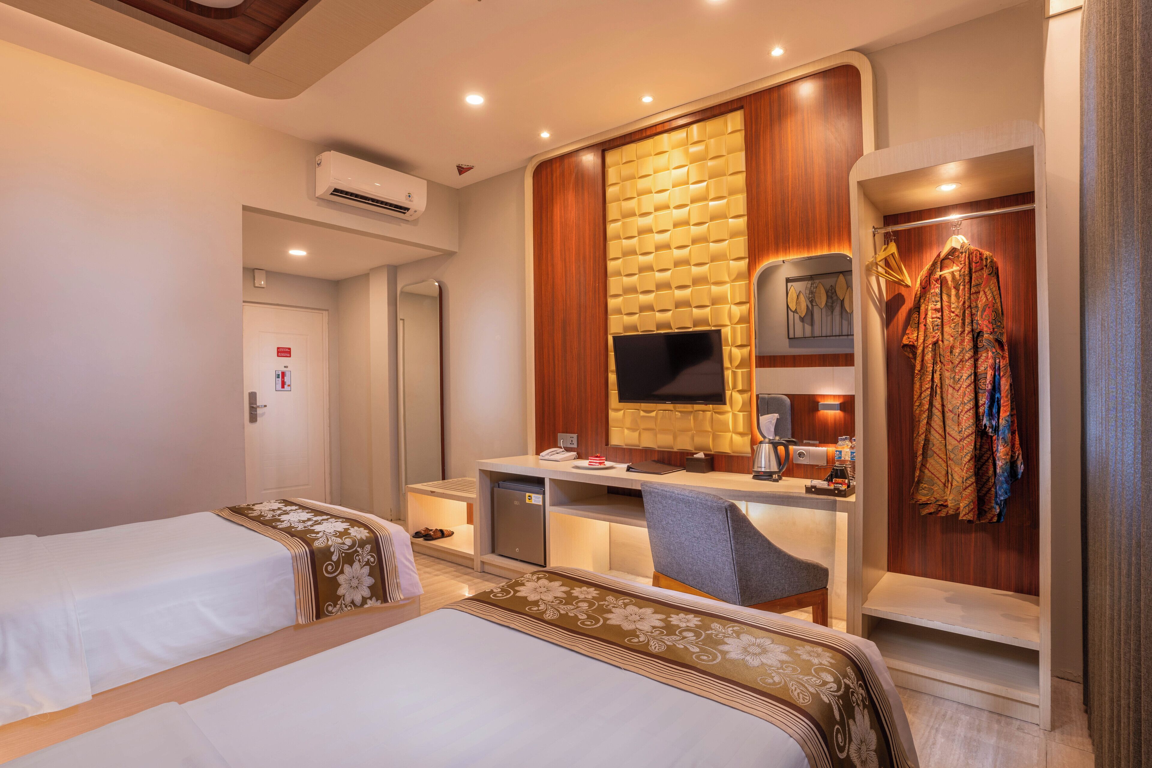 A hotel room with two beds, a desk with a flat-screen TV, a wardrobe, and a small seating area.