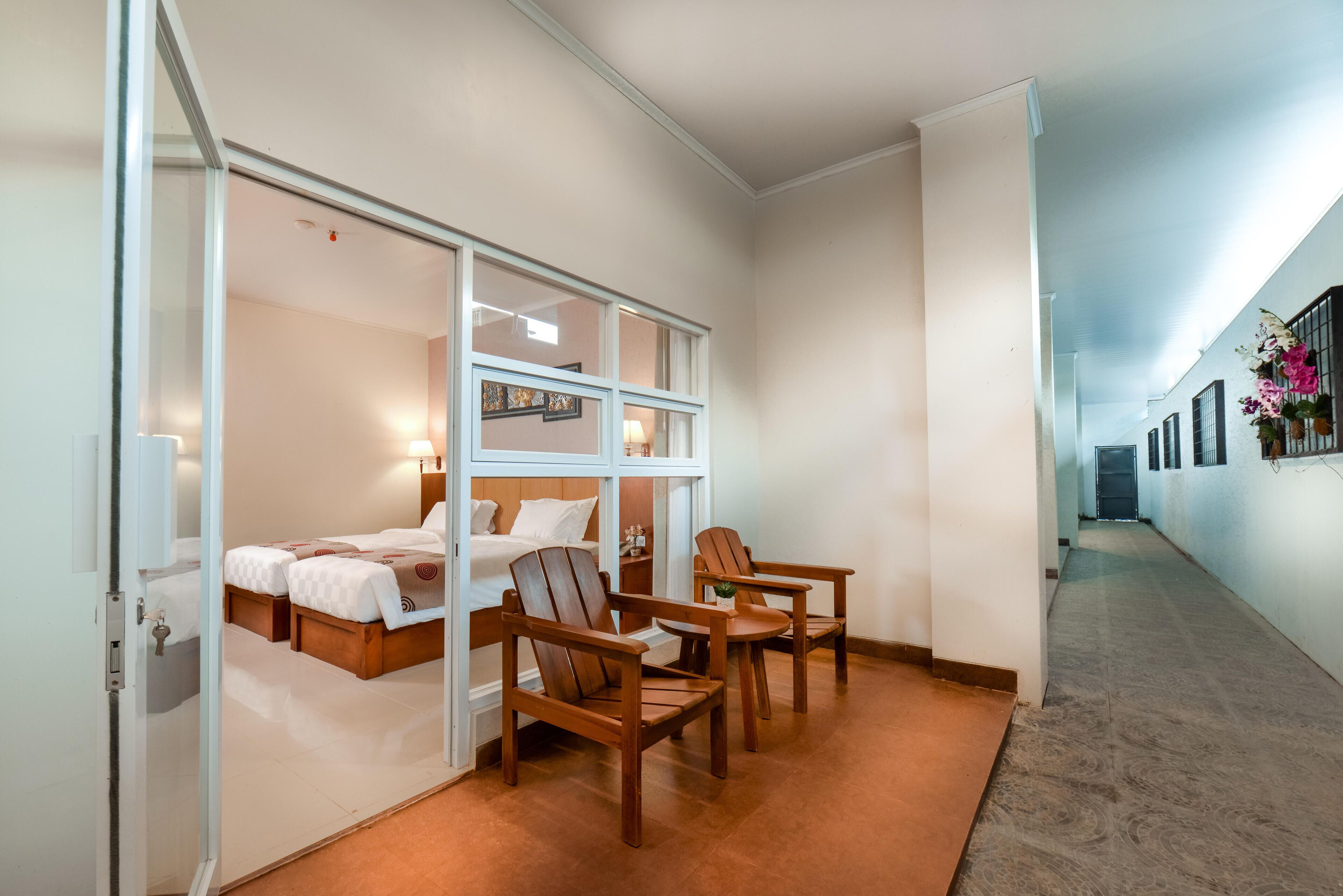 Una habitación de hotel con una cama, dos sillas de madera, una mesita y un separador de vidrio.