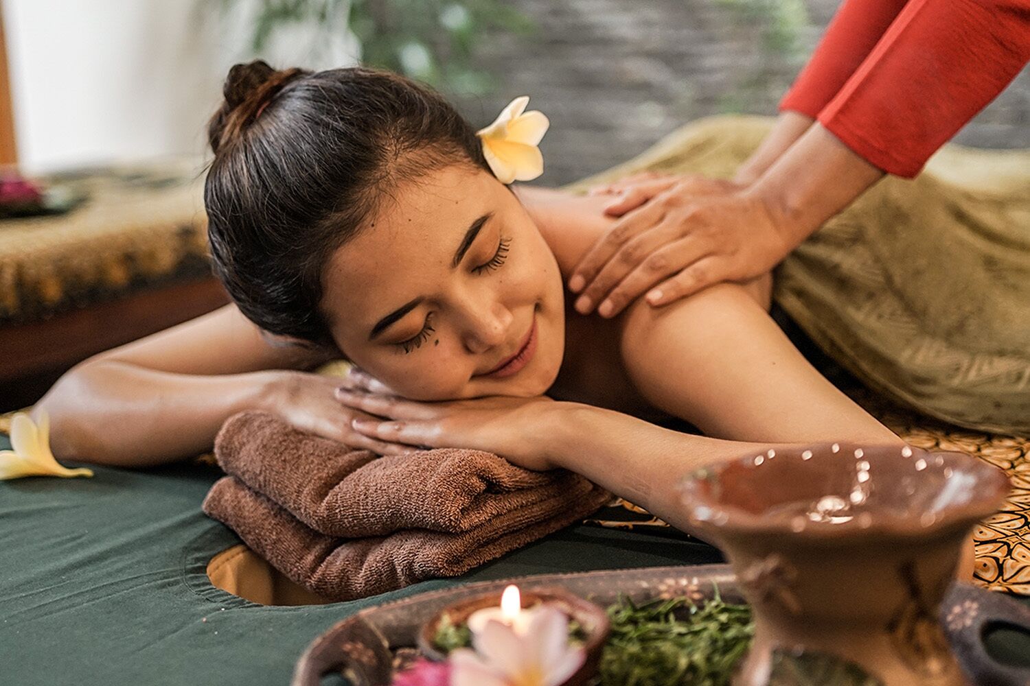 A person receiving a massage on a bed with a towel, a candle, and a small bowl of herbs.