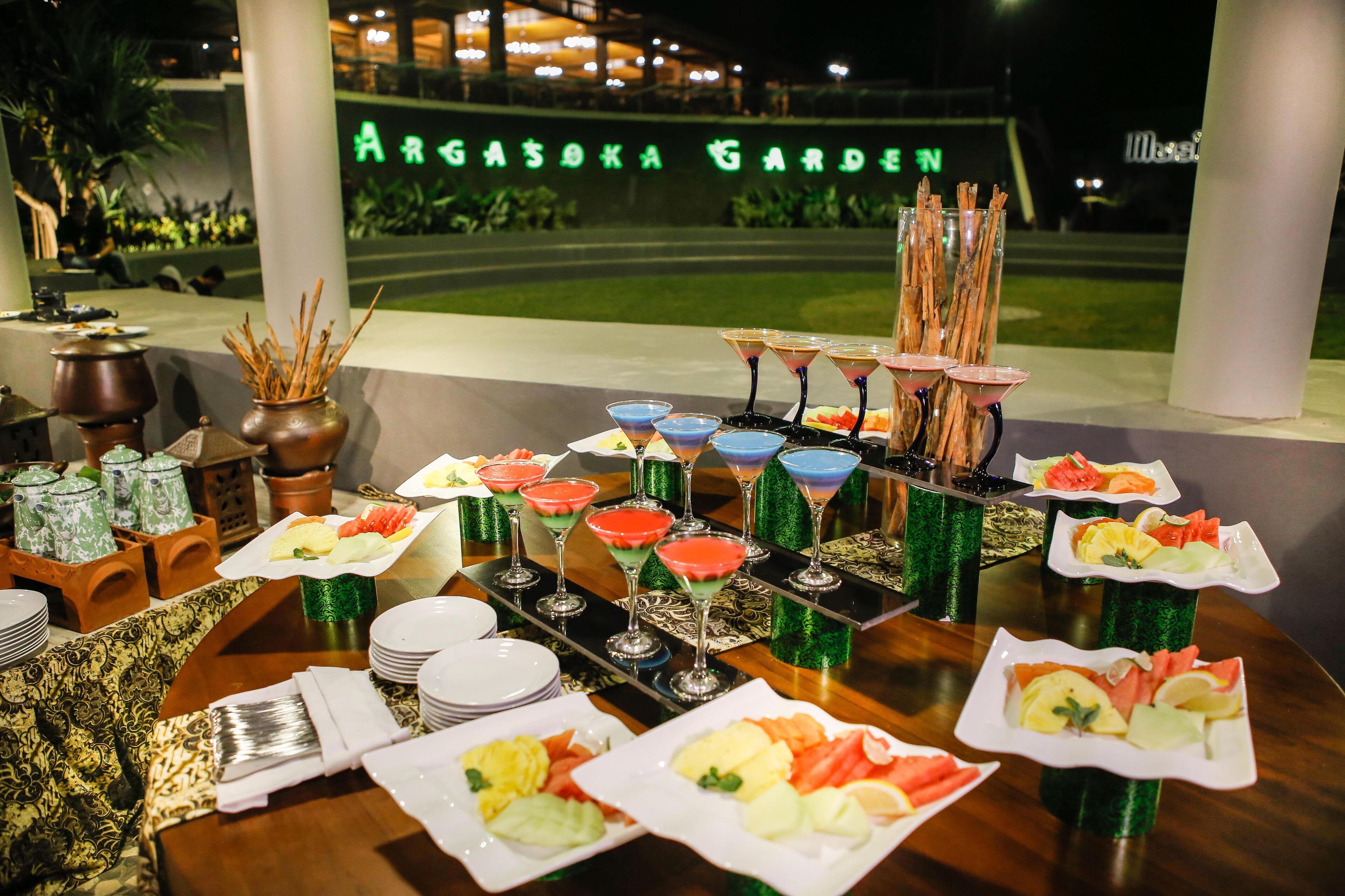 Una mesa de buffet con diversos platos y bebidas, con el jardín bien iluminado de fondo y un cartel que dice "ARGASOKA GARDEN".