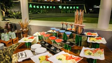 Una mesa de buffet con diversos platos y bebidas, con el jardín bien iluminado de fondo y un cartel que dice "ARGASOKA GARDEN".