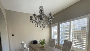 A dining room with a wooden table, four chairs, a chandelier, and large windows with blinds.