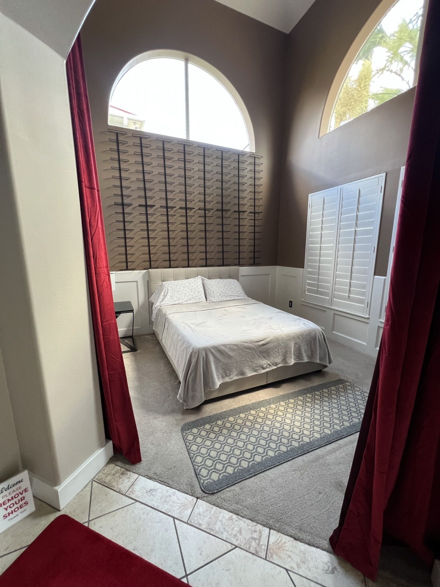 A bedroom with a bed, a window with blinds, a red curtain, a patterned rug, and a sign on the floor.