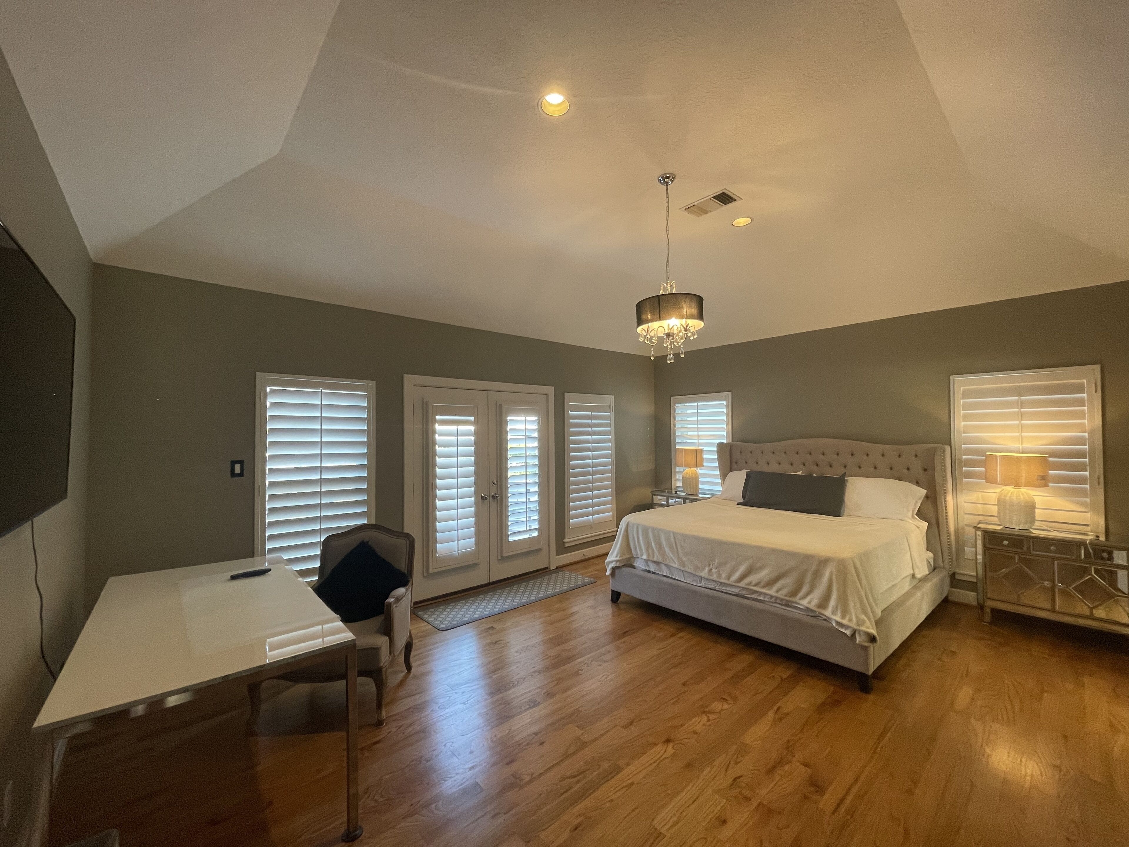 A bedroom with a bed, desk, and chair, featuring a chandelier, large windows with blinds, and a wooden floor.