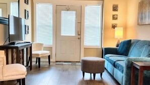 A living room with a sofa, ottoman, and a television on a stand. - Downtown Bryson City Flat - Walk to it All! (Bryson City)