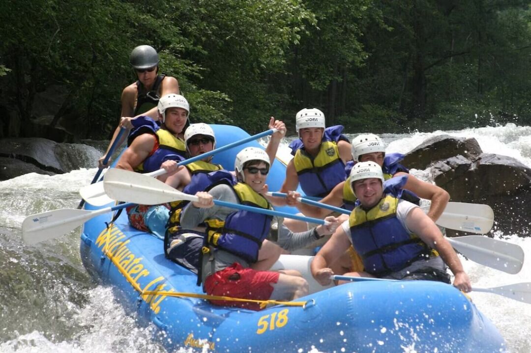 A group of people in a blue inflatable raft with the number 518, paddling on a river surrounded by trees.