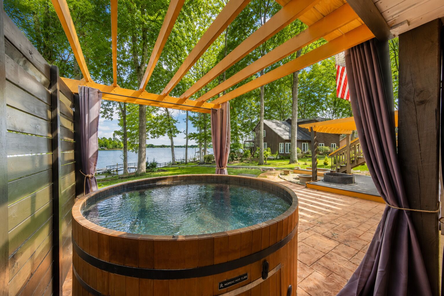 Un jacuzzi con revestimiento de madera, rodeado por una terraza de madera y un área cubierta con vista a un lago y una casa.