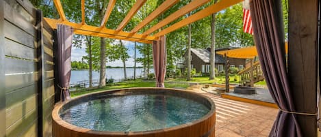 A hot tub with a wooden exterior, surrounded by a wooden deck and a covered area with a view of a lake and a house.