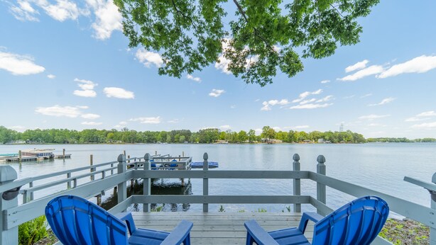 A wooden deck with two blue chairs facing a calm lake, a dock with boats, and a clear sky. - Sunny Daze by SoCharm | Hot Tub | Fire Pit | Dock | Great Views (Denver)